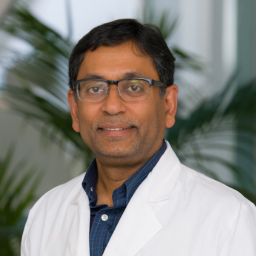 Man in white lab coat and blue shirt with a plant in the background